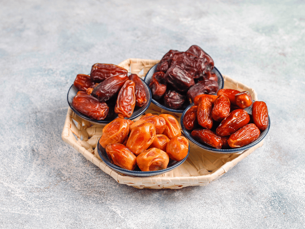 Top view of assorted date varieties, served in individual bowls on a woven tray, showcasing a range of textures and colors from golden brown to deep black, perfect for gourmet gifting or nutritional comparison.